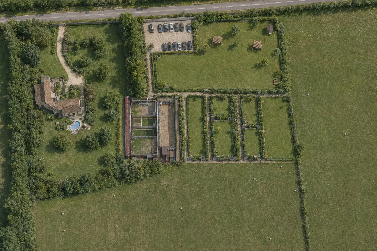 Vue aérienne du cœur du Domaine animalier des Brévonnes : la route en haut, le stationnement, les bâtiments à toiture rouge de la pension, les parcs clôturés et les prairies